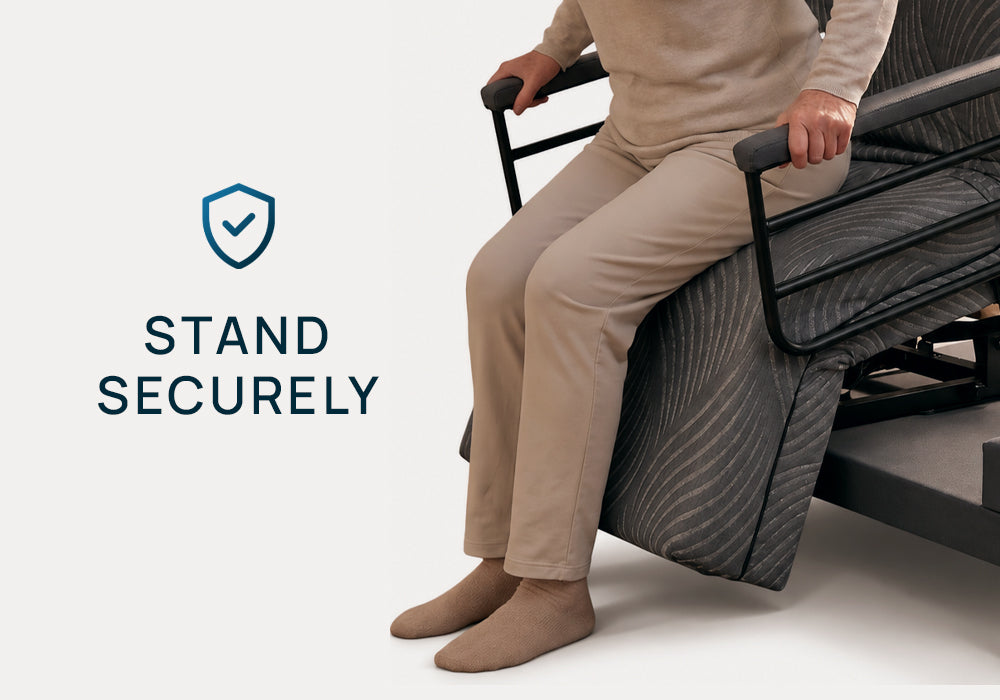 A person in beige sits on the Independence Bed holding the armrests while the bed is being pushed upward. The text 'STAND SECURELY' with a blue shield icon appears on the left side of the image.